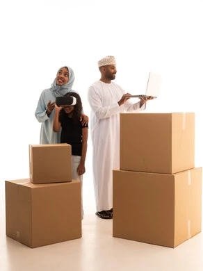 صناديق وحاويات كرتونية، استخدام تقنيات حديثة، امتلاك سكن جديد والاستقرار العائلي، بنت ترتدي نظارة الواقع الإفتراضي ثلاثية الأبعاد، الانتفال الى منزل جديد، بورتريه لرجل عربي خليجي عماني يرتدي الدشداشة و الكمة يستخدم الحاسوب المحمول برفقة زوجته و ابنته، الجسم بالطول الكامل، خلفية بيضاء