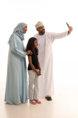 Taking a commemorative selfie via mobile phone, caring for and paying attention to children, an Omani family spending enjoyable times, gestures of happiness and joy, a portrait of an Arab Gulf Omani man wearing a dishdasha and kumma standing with his wife and daughter, enhancing social relationships, full-body shot, white background.