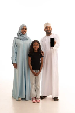 تعزيز العلاقات الاجتماعية، عرض شاشة سوداء عبر الهاتف المحمول ، الاشارة الى شيء ما، عائلة عمانية تقضي اوقات ممتعة، النظر إلى الكاميرا بايماءات السعادة والسرور، بورتريه لرجل عربي خليجي عماني يرتدي الدشداشة والكمة يقف مع زوجته وابنته، الجسم بالطول الكامل، خلفية بيضاء