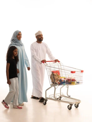 شراء المستلزمات والحاجيات الضرورية، الاستفادة من الخصومات على المنتجات، الاستمتاع بالتسوق في المولات والمراكز التجارية، بورتريه لرجل عربي خليجي عماني يرتدي الدشداشة و الكمة يجرعربة التسوق برفقة زوجته و ابنته، الجسم بالطول الكامل، خلفية بيضاء