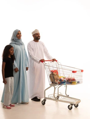 الاستفادة من الخصومات على المنتجات، الاستمتاع بالتسوق في المولات والمراكز التجارية، شراء المستلزمات والحاجيات الضرورية، بورتريه لرجل عربي خليجي عماني يرتدي الدشداشة و الكمة يجرعربة التسوق برفقة زوجته و ابنته، الجسم بالطول الكامل، خلفية بيضاء