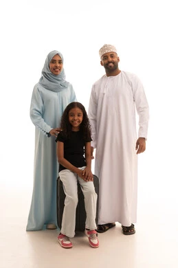 Looking at the camera with gestures of happiness and joy, a portrait of an Arab Gulf Omani woman wearing an abaya and hijab standing with her husband and daughter next to a suitcase, preparing to spend the summer vacation and go on a trip, a family atmosphere filled with love, full-body shot, white background.