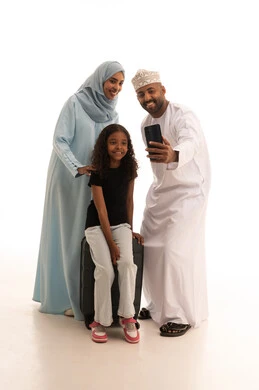 التقاط صورة سيلفي تذكارية عبر الهاتف المحمول، بورتريه لرجل عربي خليجي عماني يرتدي الدشداشة والكمة يقف مع زوجته ابنته وبجانبهم حقيبة الأمتعة، الاستعداد لقضاء الإجازة الصيفية والذهاب في رحلة سفر، الجسم بالطول الكامل، خلفية بيضاء