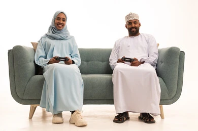 الاستمتاع والتسلية في العطلة ، استخدام التكنولوجيا الحديثة، زوجان عربيان عمانيان خليجيان يجلسان على كنبه مريحه في غرفه المعيشه يمسكان عصا التحكم ، إيماءات الاندماج و التركيز ، خلفية بيضاء