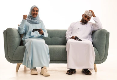 تعابير الحزن للخسارة، نشاط ترفيهي منزلي، استخدام جهاز تكنولوجي حديث، لعب ألعاب الفيديو في المنزل، بورتريه لامراة عربيه خليجيه عمانية ترتدي العباءة و الحجاب تجلس على الاريكة بجانب زوجها تحمل بيديها عصا التحكم، رجل يضع يدية على رأسة بايماءات الاحباط، خلفية بيضاء