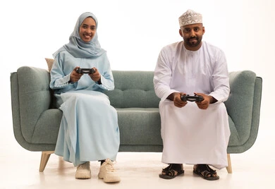الاستمتاع والتسلية في العطلة، نشاط ترفيهي منزلي، استخدام جهاز تكنولوجي حديث، لعب ألعاب الفيديو في المنزل، بورتريه لرجل عربي خليجي عماني يرتدي الدشداشة والكمة يجلس على الكنبة في غرفة المعيشة بجانب زوجته يحمل بيده عصا التحكم، خلفية بيضاء