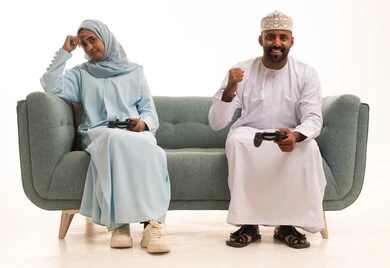 Home entertainment activity, expressions of sadness for loss, use of modern technology device, playing video games at home, portrait of an Arab Gulf Omani man wearing a dishdasha and kumma sitting on the couch next to his wife holding a game controller, a woman putting her hand on her head with gestures of frustration, white background.