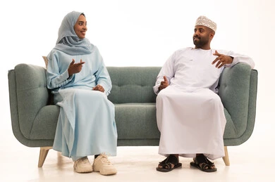 تبادل الاحاديث والاخبار بين افراد العائلة، المحافظة على الترابط الأسري، مفهوم الأمان والإستقرار العائلي، بورتريه لزوجين عربيي خليجيين عمانيين يجلسان على اريكة مريحة في غرفة المعيشة، خلفية بيضاء