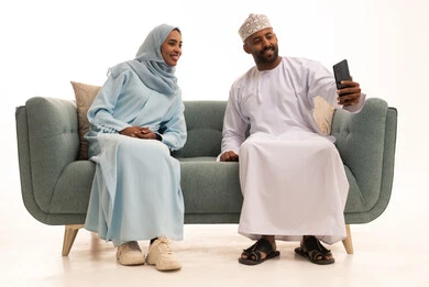 توثيق اللحظات العائلية السعيدة، بورتريه لزوجان عربيان خليجيان عمانيان يرتديان الزي التقليدي يجلسان على الاريكة يلتقطان صور سيلفي باستخدام الهاتف المحمول، قضاء اوقات ممتعة في عطلة نهاية الاسبوع، المودة والمحبة بين أفراد الأسرة، خلفية بيضاء