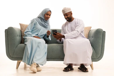 Watching movies and series on mobile, following news on social media, strengthening marital relationships and bonds of love and affection, a portrait of an Arab Gulf couple from Oman wearing traditional attire sitting on a sofa enjoying using a mobile phone, white background.