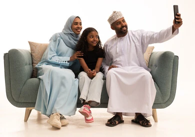 Capturing a memorable family selfie via mobile phone, the joy of children spending quality time with their parents, a portrait of a smiling Omani Arab Gulf family sitting on the couch documenting enjoyable moments with gestures of happiness and joy, a family gathering in the living room and building a growing Omani community, white background.