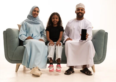 عرض شاشة سوداء فارغه عبر الجوال، مفهوم الاستقرار العائلي، استخدام تطبيقات الهاتف المحمول، بورتريه لعائلة عربية خليجية عمانية تجلس على الاريكه وتنظر الى الكاميرا بإيماءات السعادة، خلفيه بيضاء