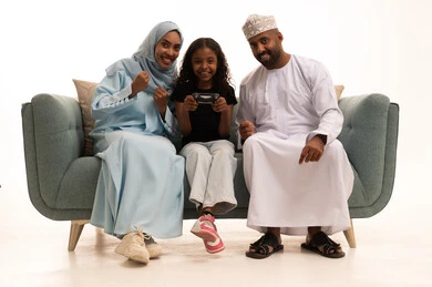 Sharing fun with the family, playing entertaining video games, using modern technology, a portrait of a Gulf Arab Omani girl sitting on the couch with her parents holding a game controller in her hand, spending enjoyable time with the children, happy family atmosphere, white background.