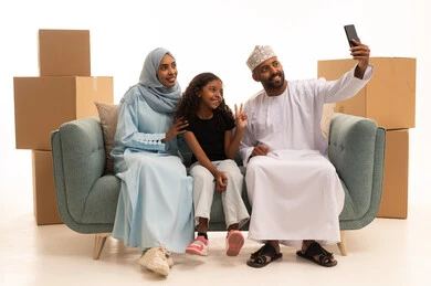 التقاط صور عائلية تذكارية من خلال الجوال، بورتريه لعائلة خليجية عمانية في غرفة المعيشة، بث فيديو مباشر، صناديق وحاويات كرتونية، مفهموم امتلاك سكن جديد والاستقرار العائلي، خلفية بيضاء