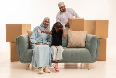 المتعة و التسلية بالمشاهدة عبر نظارة الواقع الافتراضي، بورتريه لبنت عربيه خليجيه عمانية ترتدي الزي الكاجوال تضع نظارة الفي ار و تجلس على الاريكة برفقة والديها ترفع يدها باندماج, صناديق وحاويات كرتونية، خلفية بيضاء