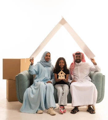 صناديق وحاويات كرتونية، زوجان عربيان خليجيان سعوديان يجلسان على الايكة برفقه ابنتهم يحملان لوح ابيض فوق راسهم، بورتريه لعائلة عربية خليجية سعودية، بنت تمسك بيديها مجسم بيت صغير، ايماءات وجه ويدين تدل على السعادة، مفهوم امتلاك سكن جديد والاستقرار العائلي، خلفية بيضاء