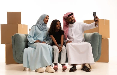 The concept of family safety and stability, boxes and cardboard containers, documenting happy family moments, a portrait of a Saudi Arabian Gulf man wearing traditional attire and a ghutrah sitting on the sofa with his wife and daughter taking a commemorative photo with a mobile phone, maintaining family ties, white background.