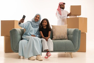 صناديق وحاويات كرتونية، مفهوم الأمان والإستقرار العائلي، توثيق اللحظات العائلية السعيدة، بورتريه لام عربيه خليجيه سعودية ترتدي العباءة و الحجاب تجلس على الاريكة برفقة ابنتها تلتقط صورة تذكارية من خلال الهاتف المحمول، المحافظة على الترابط الأسري، خلفية بيضاء