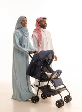 Going on family outings and trips, a Saudi family spending enjoyable times, caring for and paying attention to the children, the importance of parents and their essential role in raising and nurturing children, a portrait of a Saudi Gulf Arab man wearing traditional attire and a ghutrah standing with his wife and pushing a stroller, white background.