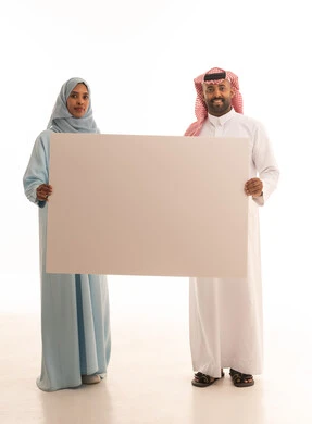 إعلانات وعروض تسويقية، استخدام لوحات توضيحية ووسائل تعليمية، بورتريه لرجل عربي خليجي سعودي يرتدي  الثوب التقليدي و الشماغ يحمل بيده لوح ابيض كبير (موك اب فارغ) برفقة زوجته ، ايماءات السعادة والسرور، الطول الكامل للجسم، خلفية بيضاء
