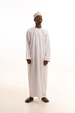 Standing upright and steady, paying attention to external appearance and elegance, the concept of masculinity and self-confidence, a portrait of a young Arab Gulf Omani man with a dark complexion wearing a dishdasha and kumma, looking with expressions of joy, full-body image, white background.