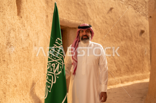 إحياء ذكرى اليوم الوطني السعودي 23 سبتمبر, إيماءات الفخر و السرور بالانتماء للوطن, رجل عربي خليجي سعودي مبتسم يرتدي الشماغ و الثوب التقليدي يقف في بيت طيني ويحمل بيده علم المملكة، يوم بدينا 1727 م، فن العمارة القديم وبناء البيوت الطينية، يوم العلم 11 مارس