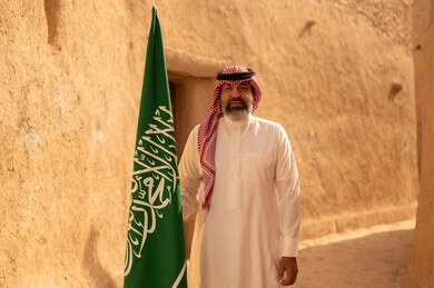 إحياء ذكرى اليوم الوطني السعودي 23 سبتمبر, إيماءات الفخر و السرور بالانتماء للوطن, رجل عربي خليجي سعودي مبتسم يرتدي الشماغ و الثوب التقليدي يقف في بيت طيني ويحمل بيده علم المملكة، يوم بدينا 1727 م، فن العمارة القديم وبناء البيوت الطينية، يوم العلم 11 مارس