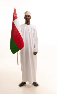 مفهوم حب الوطن ، تقديم الولاء للراية العمانية بمشاعر العز والكرامة، الفخر والاعتزاز بالانتماء لسلطنة عمان، بورتريه لشاب عربي خليجي عماني اسمر يرتدي الدشداشة والكمه التقليدية يمسك العلم العماني بيده، الجسم بالطول الكامل، خلفية بيضاء