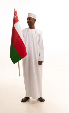 Expressing loyalty to the Omani flag with feelings of pride and dignity, pride and honor in belonging to the Sultanate of Oman, a portrait of a young Arab Gulf Omani man with a dark complexion wearing a traditional dishdasha and kumma holding the Omani flag in his hand, full-body, white background.