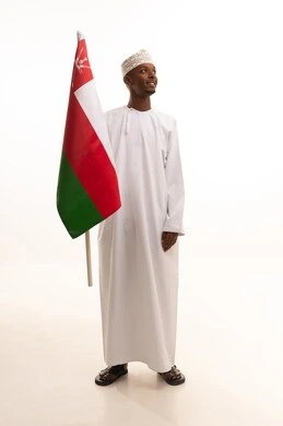 Expressing loyalty to the Omani flag with feelings of pride and dignity, pride and honor in belonging to the Sultanate of Oman, a portrait of a young Arab Gulf Omani man with a dark complexion wearing a traditional dishdasha and kumma holding the Omani flag in his hand, full-body, white background.
