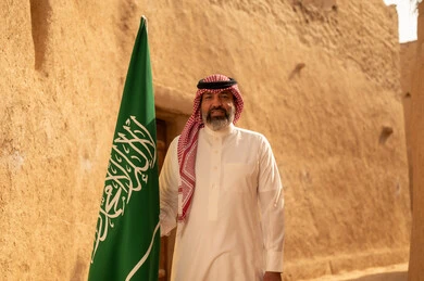 إيماءات الفخر و السرور بالانتماء للوطن, رجل عربي خليجي سعودي مبتسم يرتدي الشماغ و الثوب التقليدي يقف في بيت طيني ويحمل بيده علم المملكة، يوم بدينا 1727 م، فن العمارة القديم وبناء البيوت الطينية، يوم العلم 11 مارس، إحياء ذكرى اليوم الوطني السعودي 23 سبتمبر