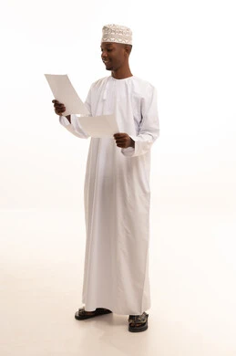 Reviewing notes, preparing work-related reports, a portrait of a young Arab Gulf Omani man with a dark complexion wearing a dishdasha and a kumma, holding a set of white papers in his hand, reviewing and following up on office plans and projects with focused gestures, white background.