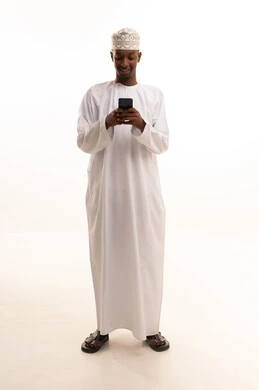 Making a phone call, using modern technologies, browsing social media sites via mobile, completing work tasks remotely, a portrait of a young Arab Gulf Omani man with a dark complexion wearing a dishdasha and kumma using a mobile phone, white background.