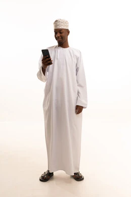 تصفح مواقع التواصل الاجتماعي عبر الجوال، اجراء مكالمة هاتفية ، استخدام التقنيات الحديثة،  إنجاز مهام العمل عن بعد، بورتريه لشاب عربي خليجي عماني اسمر يرتدي الدشداشة والكمة  يقوم باستخدام الهاتف المحمول، خلفية بيضاء