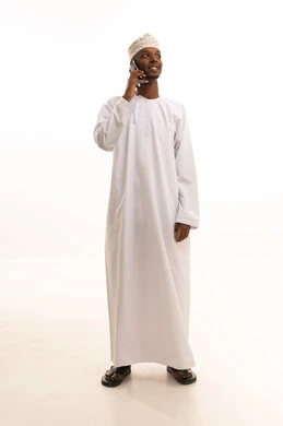 Making a phone call, using modern technologies, browsing social media sites via mobile, completing work tasks remotely, a portrait of a young Arab Gulf Omani man with a dark complexion wearing a dishdasha and kumma using a mobile phone, white background.