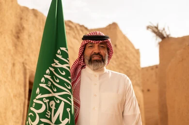 إحياء ذكرى اليوم الوطني السعودي 23 سبتمبر, إيماءات الفخر و السرور بالانتماء للوطن, رجل عربي خليجي سعودي مبتسم يرتدي الشماغ و الثوب التقليدي يقف في بيت طيني ويحمل بيده علم المملكة، يوم بدينا 1727 م، فن العمارة القديم وبناء البيوت الطينية، يوم العلم 11 مارس