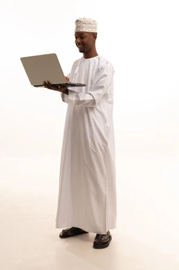 العمل في القطاع الصحي السعودي، تقديم الخدمات للمرضى بكل كفاءه، استخدام التكنولوجيا والتقنيات الحديثة، اجراء مكالمات الفيديو بتعابير الترحيب، ، مفهوم الطب والرعاية الصحية ،بورتريه لطبيب عربي خليجي عماني اسمر يرتدي الدشداشة والكمة  والمعطف الابيض يستخدم الحاسوب المحمول ، خلفية بيضاء