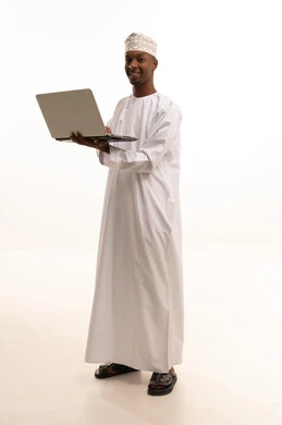 The use of technology and modern techniques, making video calls with welcoming expressions, working in the Saudi health sector, providing services to patients with high efficiency, the concept of medicine and healthcare, a portrait of a dark-skinned Arab Gulf Omani doctor wearing a dishdasha and kumma and a white coat using a laptop, white background.