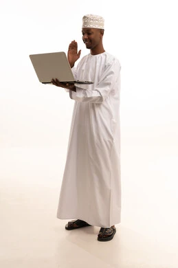 اجراء مكالمات الفيديو بتعابير الترحيب،استخدام التكنولوجيا والتقنيات الحديثة، العمل في القطاع الصحي السعودي، تقديم الخدمات للمرضى بكل كفاءه، ، مفهوم الطب والرعاية الصحية ،بورتريه لطبيب عربي خليجي عماني اسمر يرتدي الدشداشة والكمة  والمعطف الابيض يستخدم الحاسوب المحمول ، خلفية بيضاء