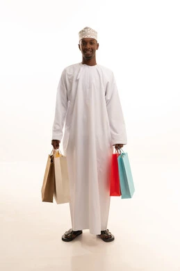 المتعة في التسوق واقتناء المستلزمات والهدايا، الاستفادة من العروضات والتخفيضات،بورتريه لشاب عربي خليجي عماني اسمر يرتدي الدشداشة والكمة يحمل بيده أكياس ورقية ملونة بإيماءات السرور والسعادة، الجسم بالطول الكامل، خلفية بيضاء