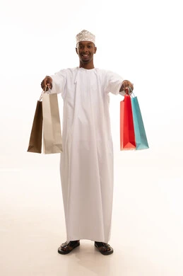 The joy of shopping and acquiring necessities and gifts, taking advantage of offers and discounts, a portrait of a young Arab Gulf Omani man with a dark complexion wearing a dishdasha and kumma, holding colorful paper bags in his hand with gestures of joy and happiness, full-body, white background.