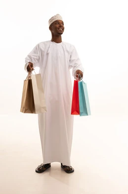 The joy of shopping and acquiring necessities and gifts, taking advantage of offers and discounts, a portrait of a young Arab Gulf Omani man with a dark complexion wearing a dishdasha and kumma, holding colorful paper bags in his hand with gestures of joy and happiness, full-body, white background.