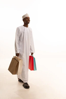 The joy of shopping and acquiring necessities and gifts, taking advantage of offers and discounts, a portrait of a young Arab Gulf Omani man with a dark complexion wearing a dishdasha and kumma, holding colorful paper bags in his hand with gestures of joy and happiness, full-body, white background.