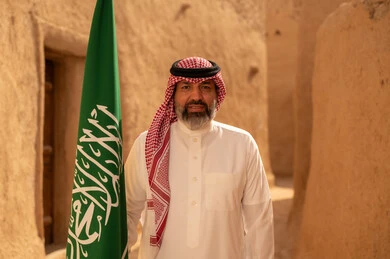 Gestures of pride and joy in belonging to the homeland, a smiling Saudi Arabian Gulf man wearing the traditional shemagh and thobe standing in a mud house and holding the flag of the Kingdom, the day we began 1727 AD, the art of ancient architecture and building mud houses, Flag Day March 11, commemorating Saudi National Day September 23