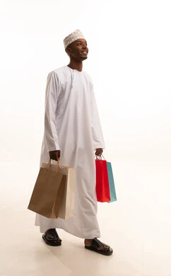 The joy of shopping and acquiring necessities and gifts, taking advantage of offers and discounts, a portrait of a young Arab Gulf Omani man with a dark complexion wearing a dishdasha and kumma, holding colorful paper bags in his hand with gestures of joy and happiness, full-body, white background.