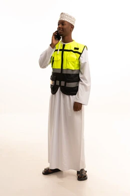 بورتريه لمهندس عربي خليجي عماني اسمر يرتدي الدشداشة والكمة  والسترة الخاصة وخوذة الحماية يقف وبيده الهاتف المحمول ، الإعمار بسواعد أبناء الوطن، مفهوم الهندسة والانشاءات المعمارية، استخدام التكنولوجيا والتقنيات الحديثة فى العمل ،المتابعة والإشراف على العمل عبر الجوال، الجسم بالطول الكامل، خلفية بيضاء