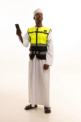 المتابعة والإشراف على العمل عبر الجوال، بورتريه لمهندس عربي خليجي عماني اسمر يرتدي الدشداشة والكمة  والسترة الخاصة وخوذة الحماية يقف وبيده الهاتف المحمول ،الإعمار بسواعد أبناء الوطن، مفهوم الهندسة والانشاءات المعمارية، استخدام التكنولوجيا والتقنيات الحديثة فى العمل ، الجسم بالطول الكامل، خلفية بيضاء