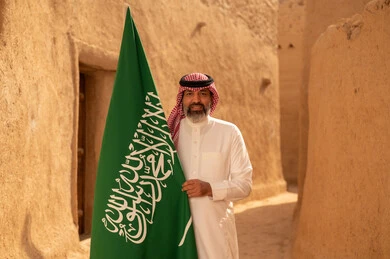 إيماءات الفخر و السرور بالانتماء للوطن, رجل عربي خليجي سعودي مبتسم يرتدي الشماغ و الثوب التقليدي يقف في بيت طيني ويحمل بيده علم المملكة، يوم بدينا 1727 م، فن العمارة القديم وبناء البيوت الطينية، يوم العلم 11 مارس، إحياء ذكرى اليوم الوطني السعودي 23 سبتمبر