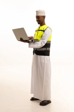المتابعة والإشراف على العمل عبر اللابتوب، استخدام التكنولوجيا والتقنيات الحديثة فى العمل ،مفهوم الهندسة والانشاءات المعمارية، بورتريه لمهندس عربي خليجي عماني اسمر يرتدي الدشداشة والكمة  والسترة الخاصة وخوذة الحماية يقف وبيده الحاسوب المحمول ، الجسم بالطول الكامل، خلفية بيضاء
