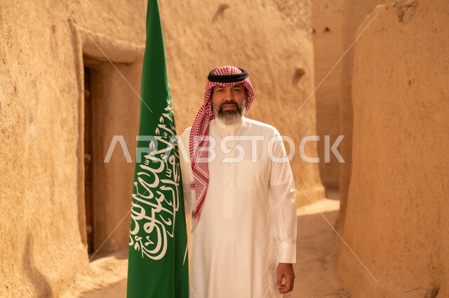 إحياء ذكرى اليوم الوطني السعودي 23 سبتمبر, إيماءات الفخر و السرور بالانتماء للوطن, رجل عربي خليجي سعودي مبتسم يرتدي الشماغ و الثوب التقليدي يقف في بيت طيني ويحمل بيده علم المملكة، يوم بدينا 1727 م، فن العمارة القديم وبناء البيوت الطينية، يوم العلم 11 مارس